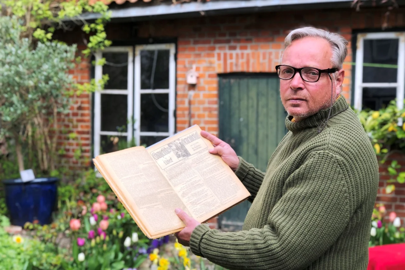 Ein Mann hält ein altes Buch aufgeschlagen in der Hand. Er zeigt die alten Seiten in Richtung Kamera. Im Hintergrund Teile eines Hauses aus Backstein mit grüner Holztür und weißen Sprossenfenstern. Frühlingsblumen blühen davor.