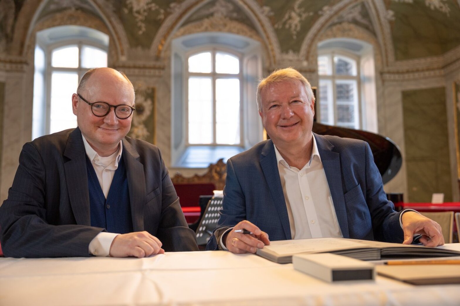 Zwei Männer sitzen hinter einem Tisch mit weißer Tischdecke und einem Dokument vor sich und lächeln in die Kamera.