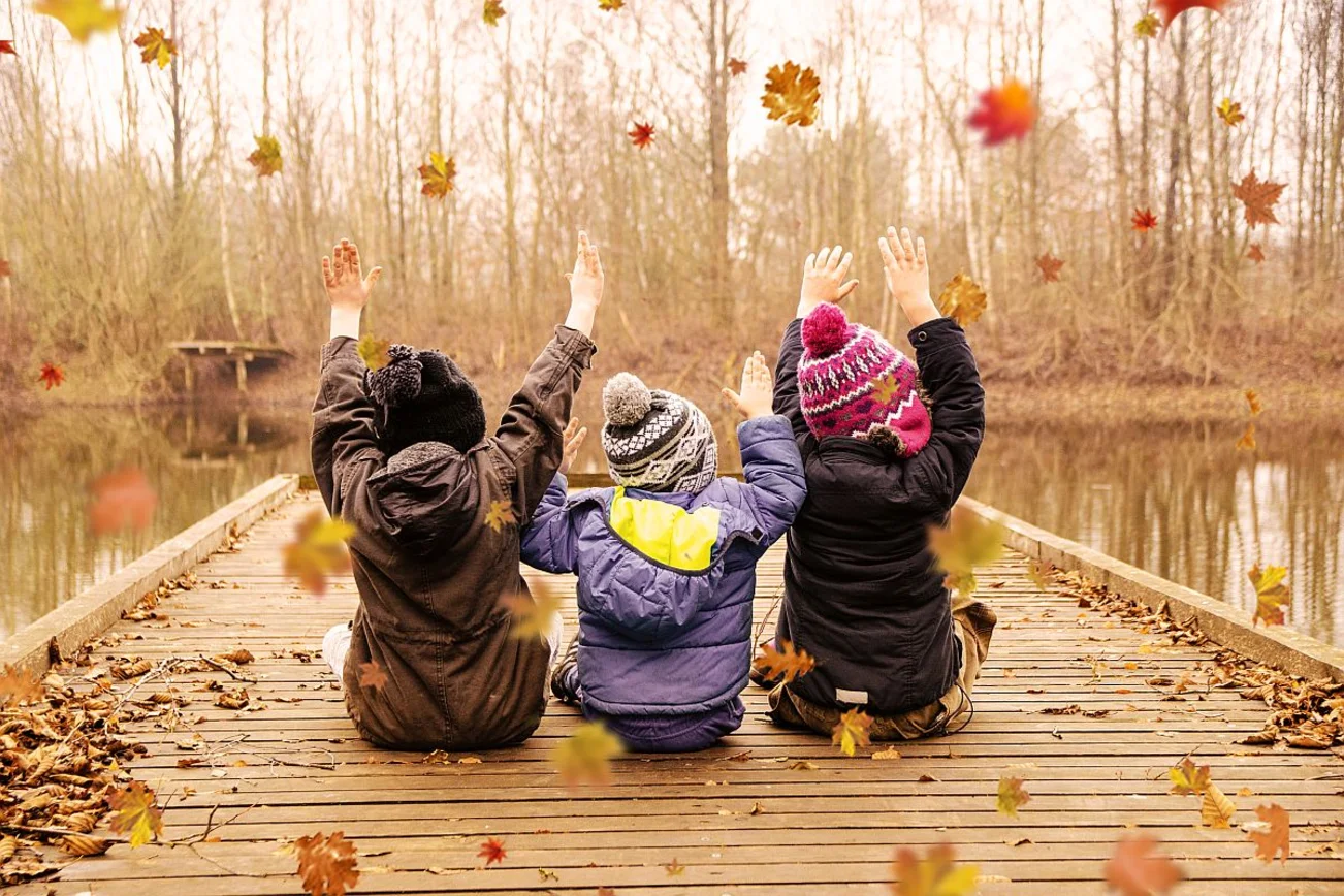 Drei Kinder sitzen auf einem Steg am See und spielen mit Blättern