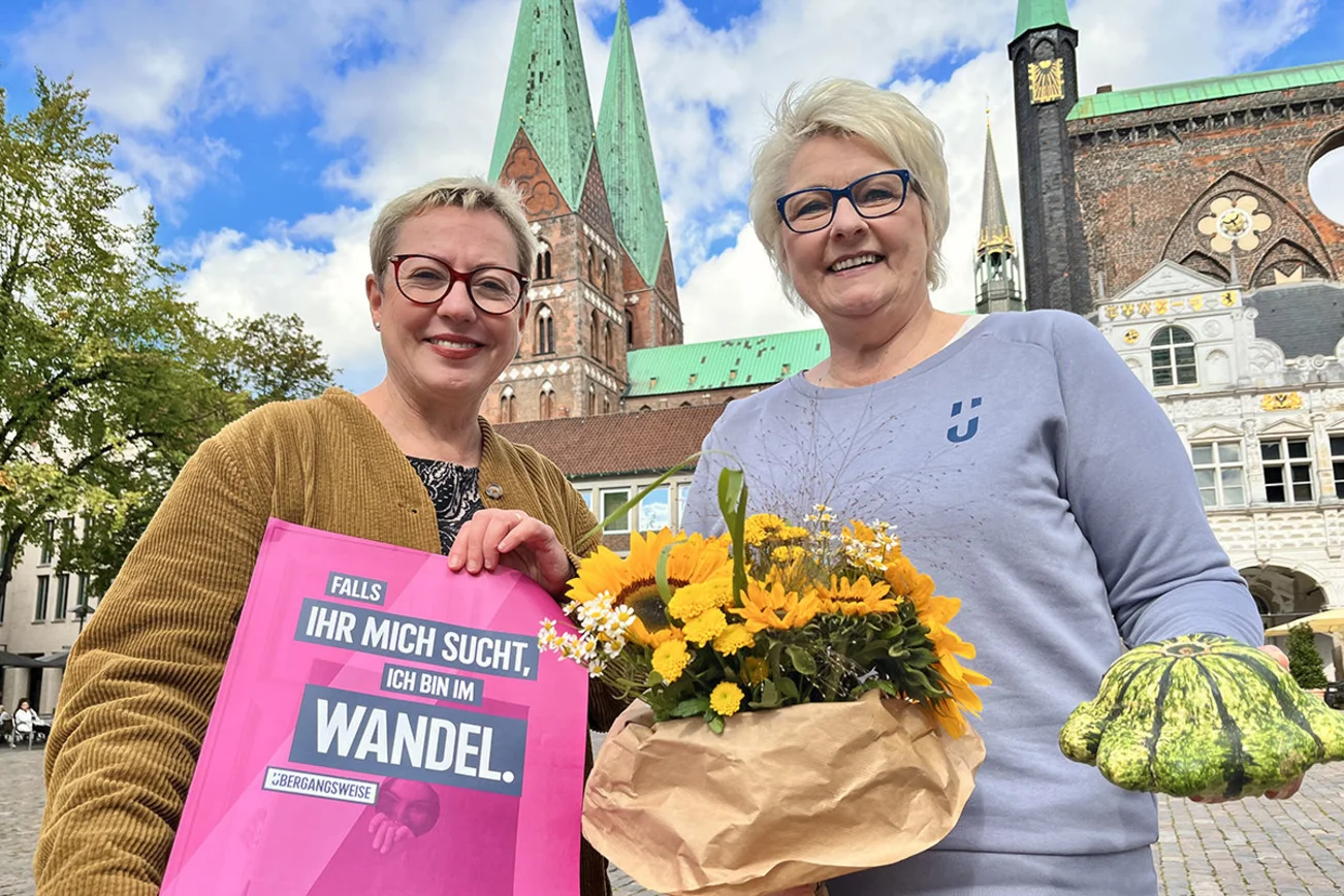 Zwei Frauen mit Blumen und Kürbis in den Händen lächeln auf dem Lübecker Marktplatz in die Kamera.