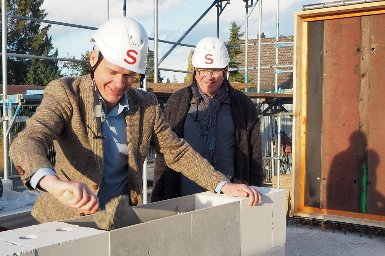 Zwei Männer mit weißen Bauhelmen stehen an deiner weißen Mauer. Der linke trägt mit einer Maurerkelle Mörtel auf.