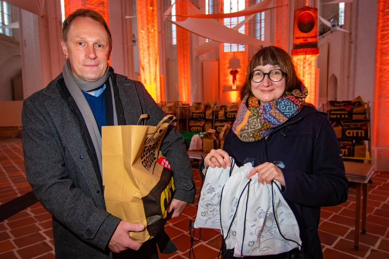 Zwei Personen stehen mit Geschenktüten in einer Kirche und lächeln in die Kamera. 