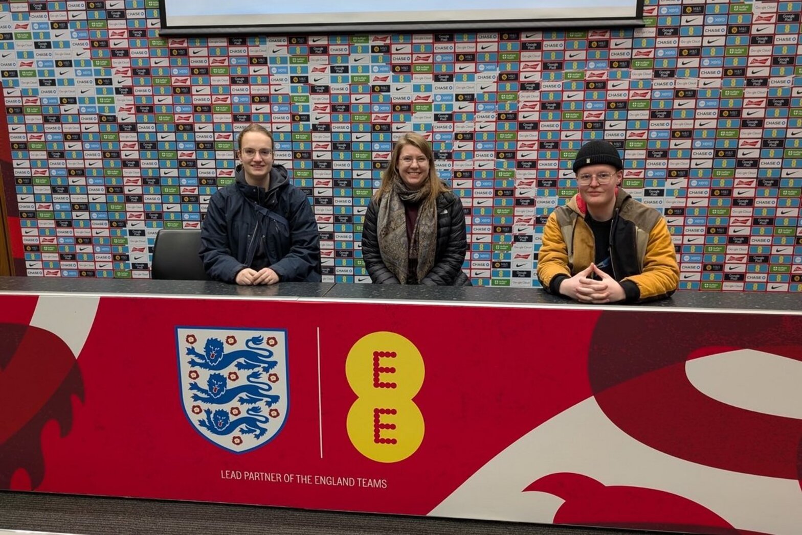 Drei junge Leute sitzen am roten Pressekonferenz-Tisch des Wembley Stadions.