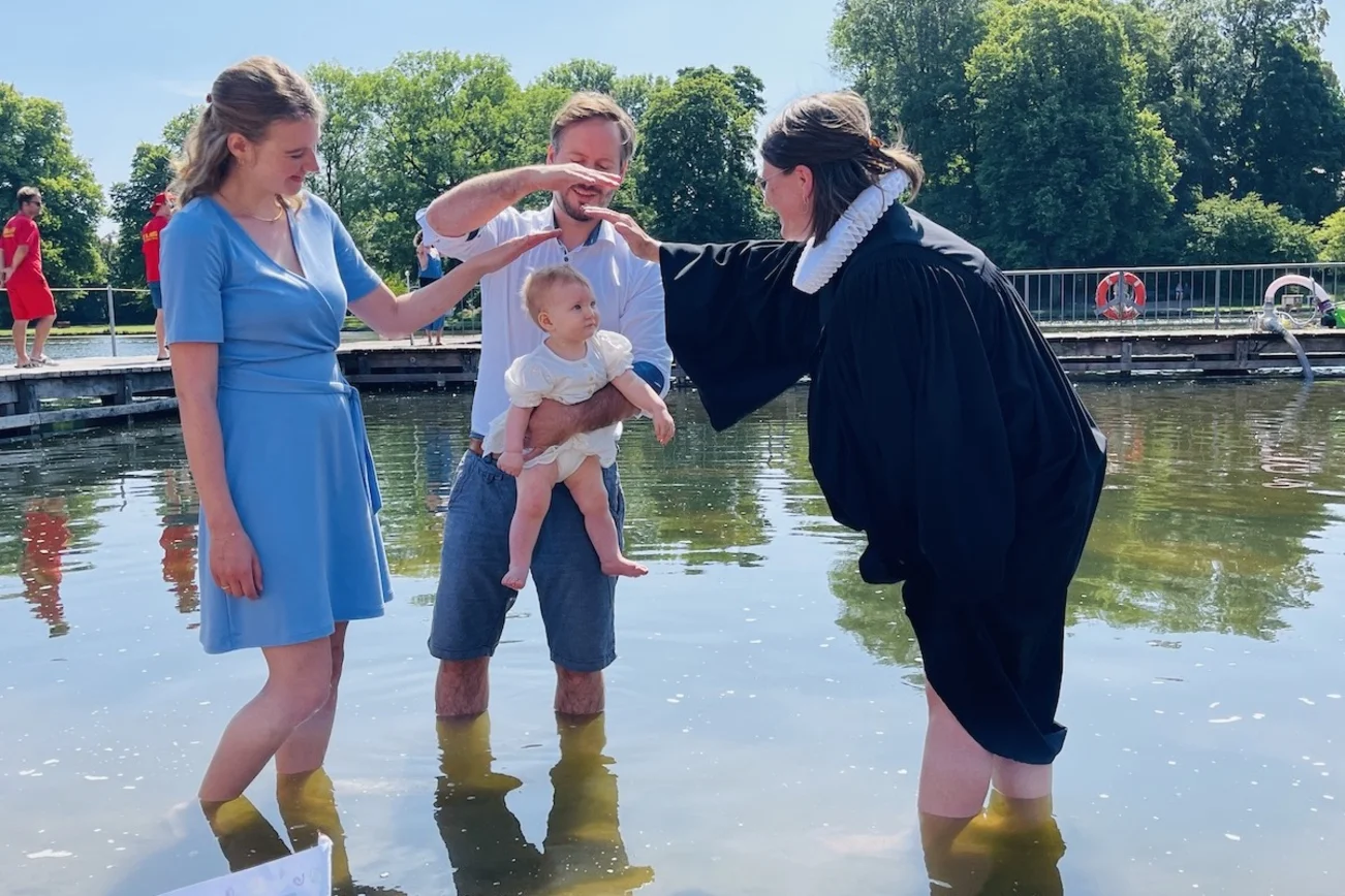 Eine Pastorin tauft ein kleines Mädchen im Krähenteich. Der Vater hält es hoch, die Mutter schaut zu freut sich. Beide Eltern halten ihre Hände als Segen über den Kopf des Kindes. 