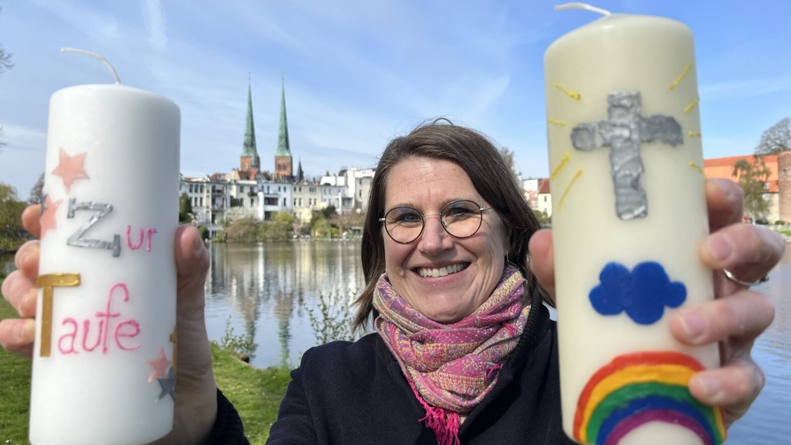 Eine Frau steht vor einem Gewässer und hält zwei Kerzen in die Kamera. 