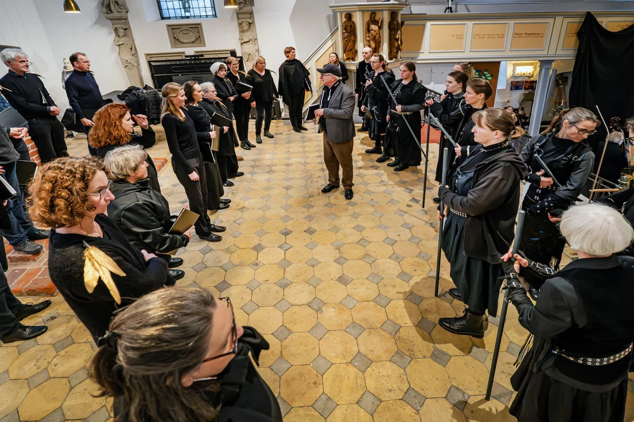 Eine Gruppe Schauspielerinnen und Schauspieler in schwarzen Uniformen steht ovalförmig in einer Kirche zusammen. 