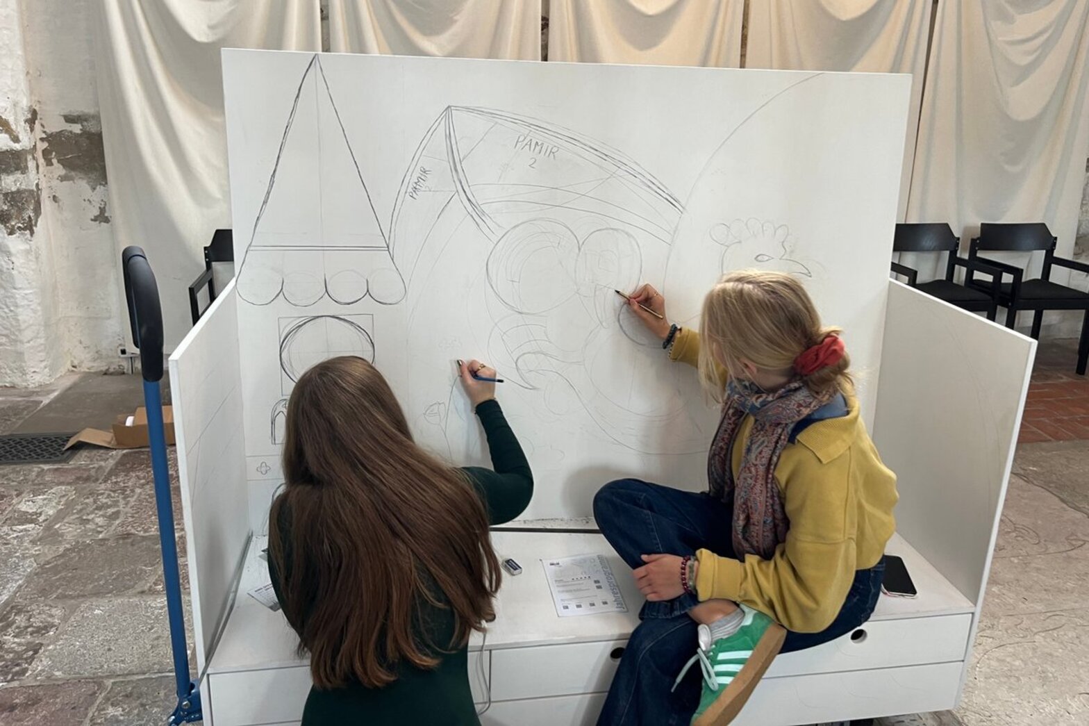 Zwei Schülerinnen skizzieren mit Bleistift auf einer Kirchenbank.