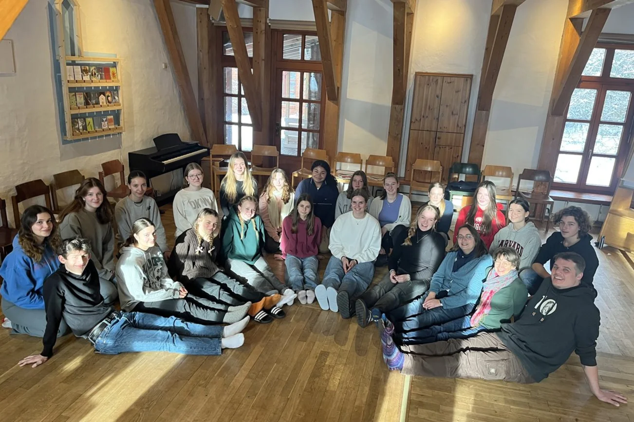Eine Gruppe Schülerinnen und Schüler sitzt im Halbkreis in einem Raum mit Holzfußboden.