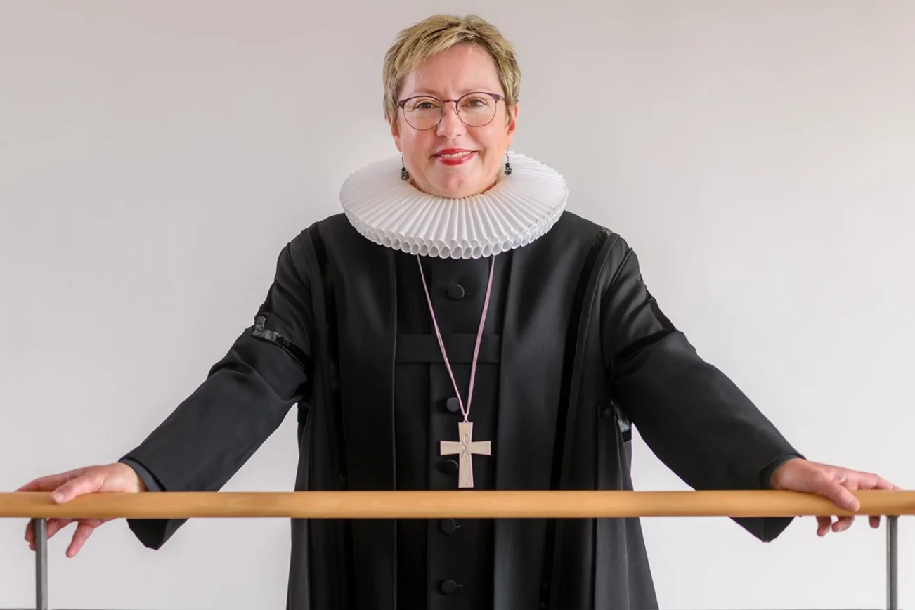 Eine Frau im Talar, mit weißem Kragen und goldenem Kreuz schaut freundlich in die Kamera.