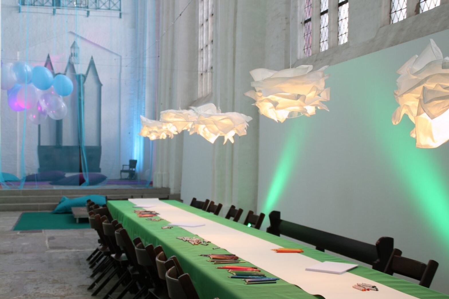 Langer Tisch mit Bastelmaterial in einer Kirche mit Wohnzimmerleuchten darüber.