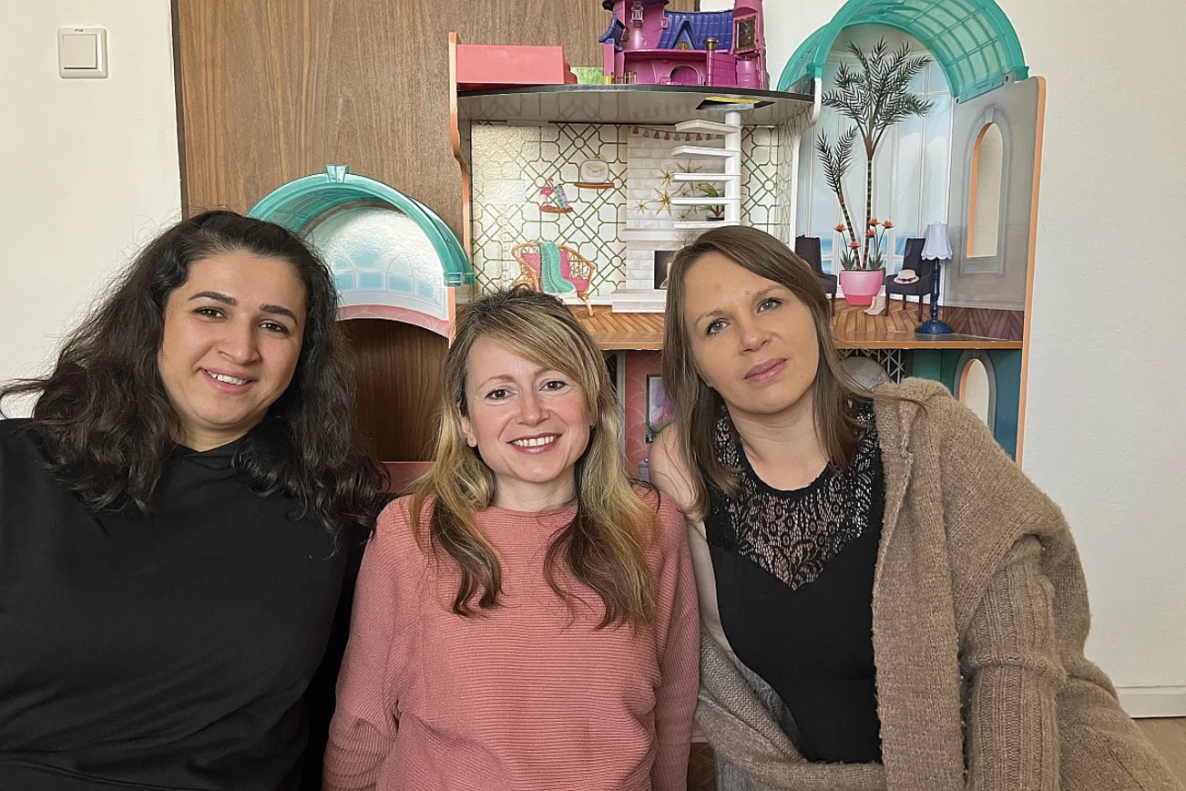 Drei Frauen sitzen vor einem großen Puppenhaus.