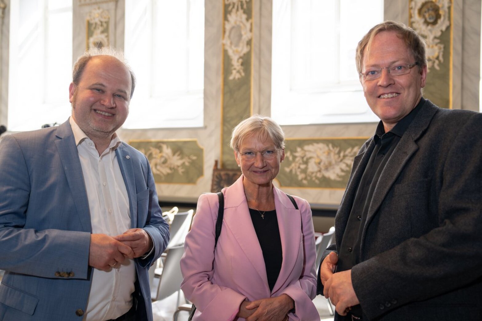 Eine Frau mit rosa Jacket steht zwischen zwei Männern mit Anzügen, alle lächeln in die Kamera.