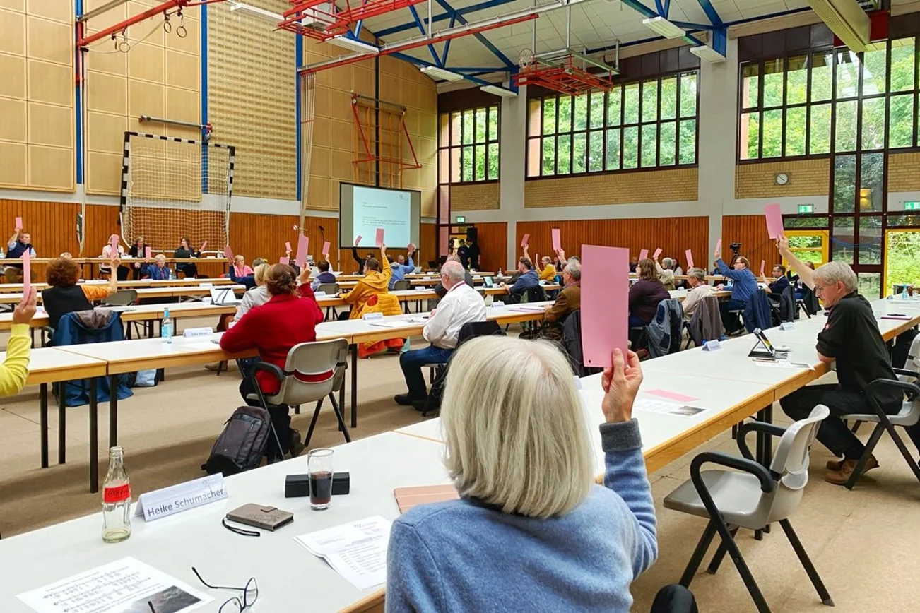 Der Blick in eine Sporthalle, die mit Tischen und Stühlen ausgestattet ist. Im Vordergrund die Rückansicht einer Frau. Sie sitzt an einem Tisch, trägt eine hellblaue Jacke und hebt eine rote Abstimmungskarte. 