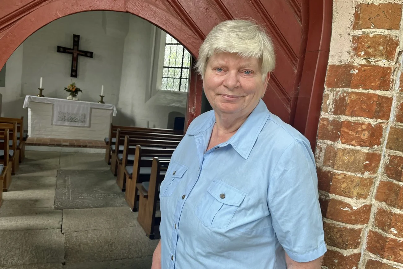 Eine Frau mit kurzen grauen Haaren steht vor dem Eingang einer Kapelle.