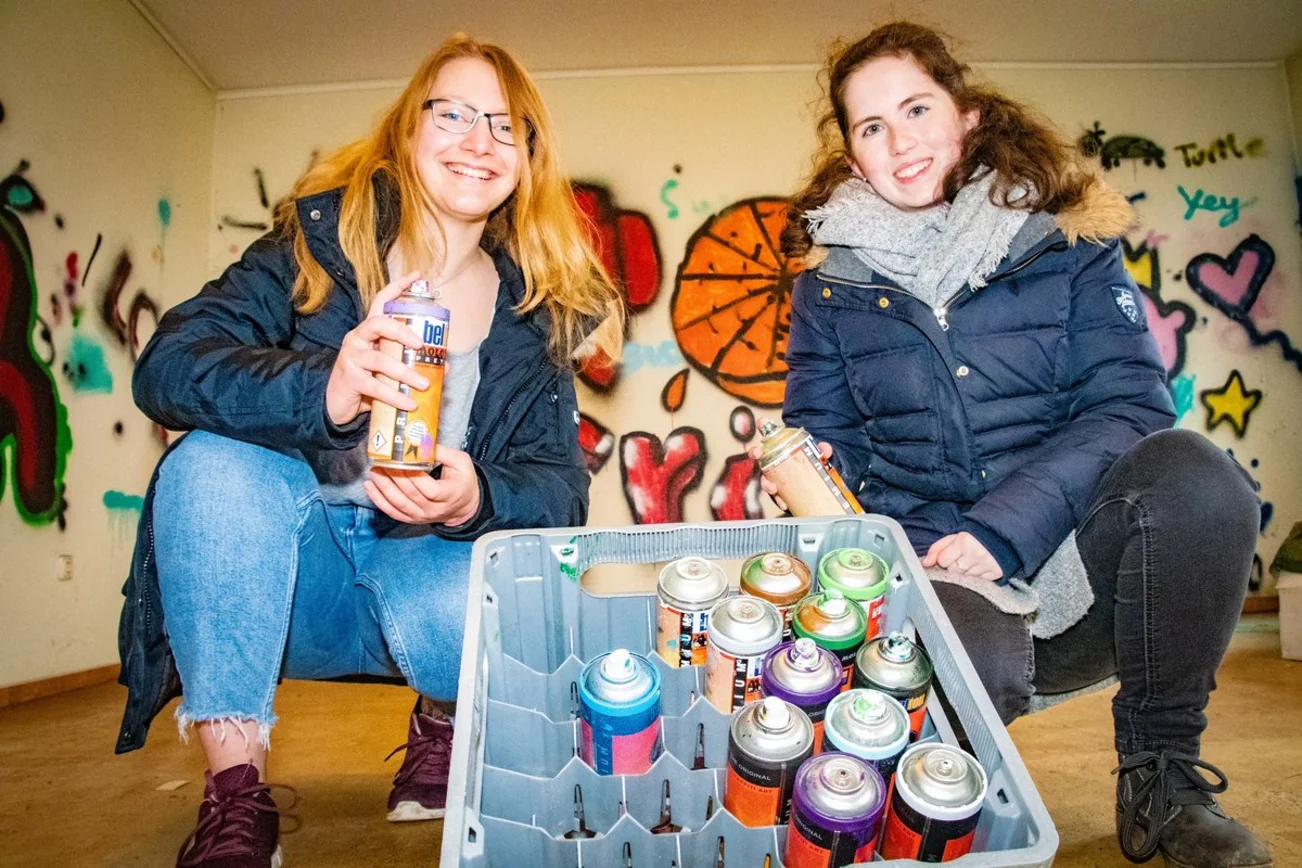 Zwei junge Frauen lächeln in die Kamera und halten Farbdosen in den Händen. 