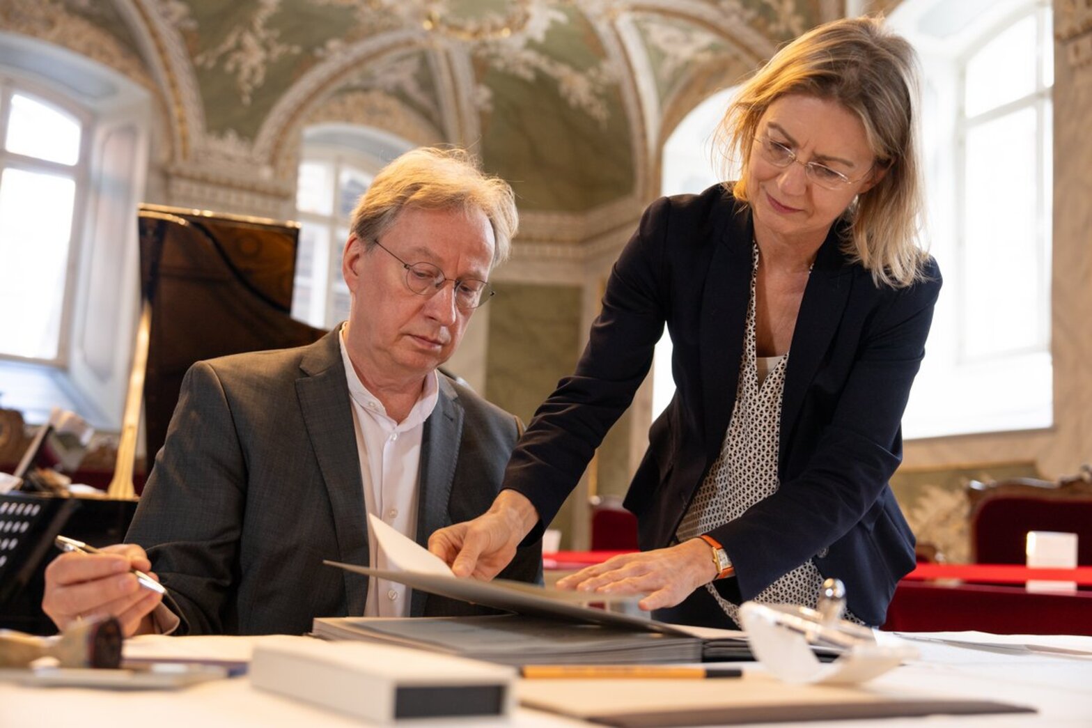 Hinter einem Tisch sitzt ein Mann mit Stift in der Hand, eine Frau rechts von ihm zeigt ihm ein Dokument.