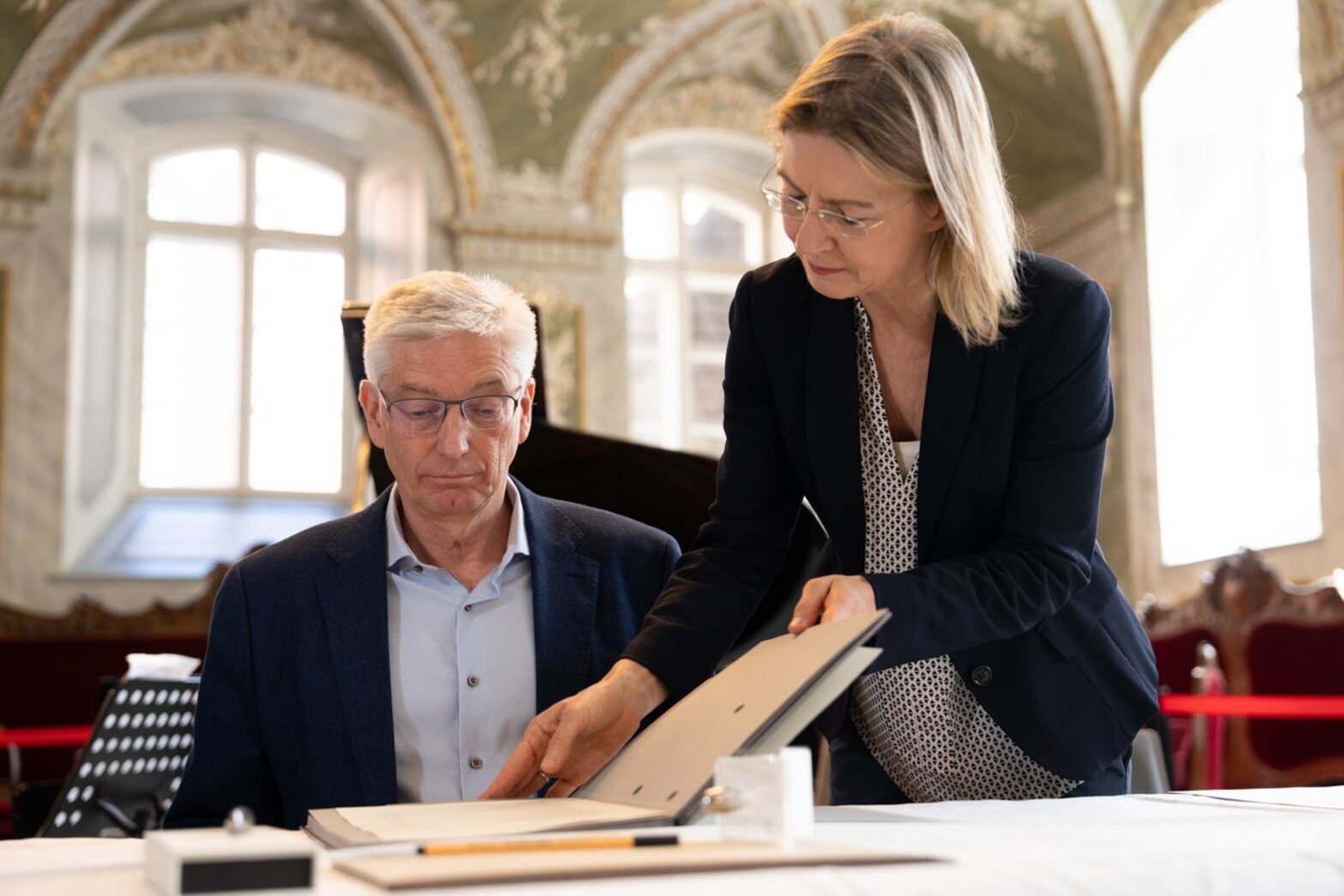 Eine Frau öffnet eine Dokumentensmappe und zeigt ein Dokument einem Mann der hinter einem Tisch sitzt. 