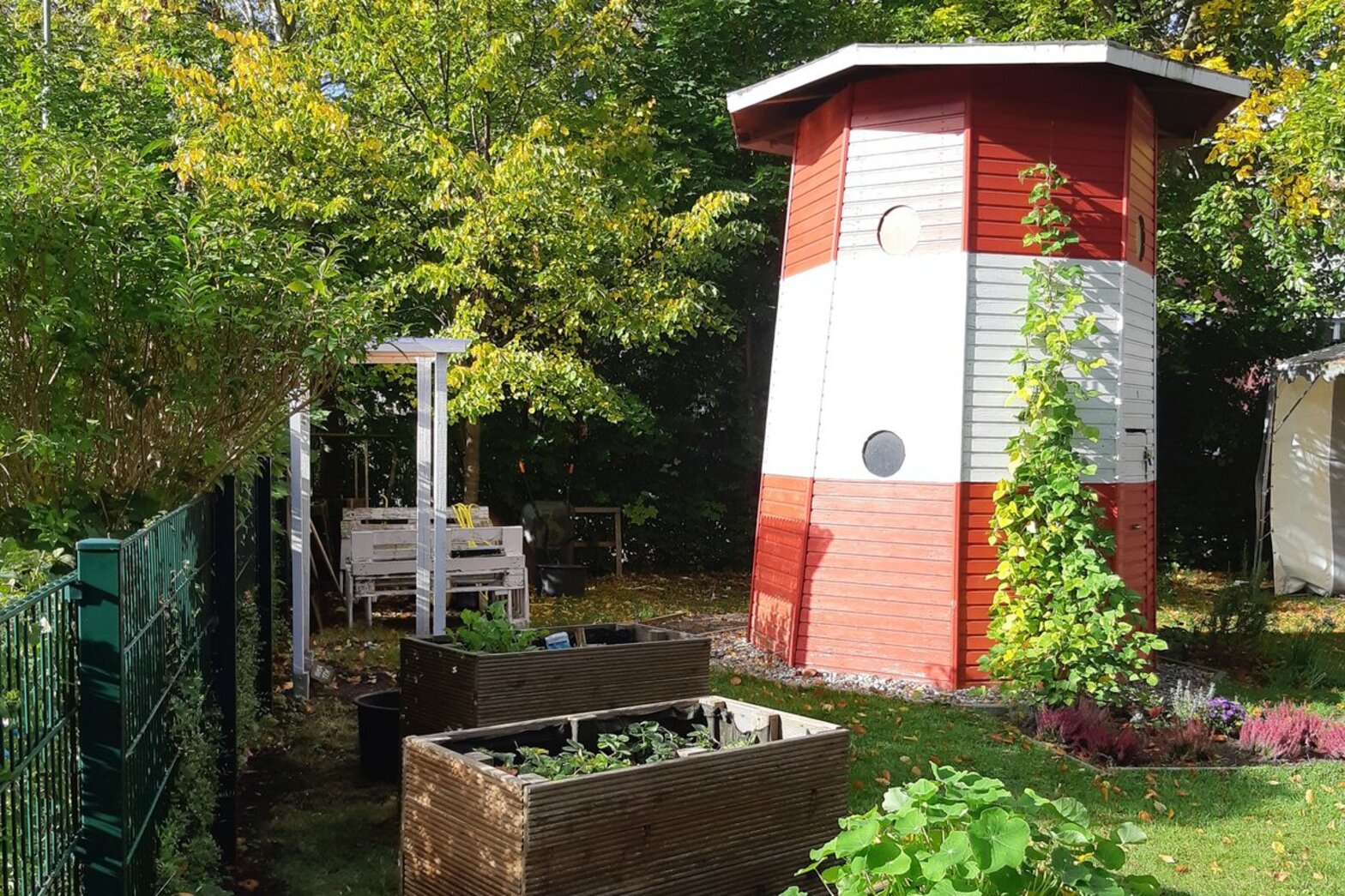 Ein breiter Leuchtturm aus Holz steht in einem Garten umgeben von Hochbeeten.