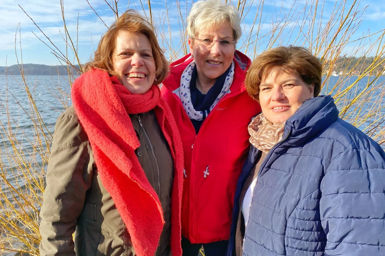 Drei Frauen stehen lächelnd am Rand eines Gewässers. 
