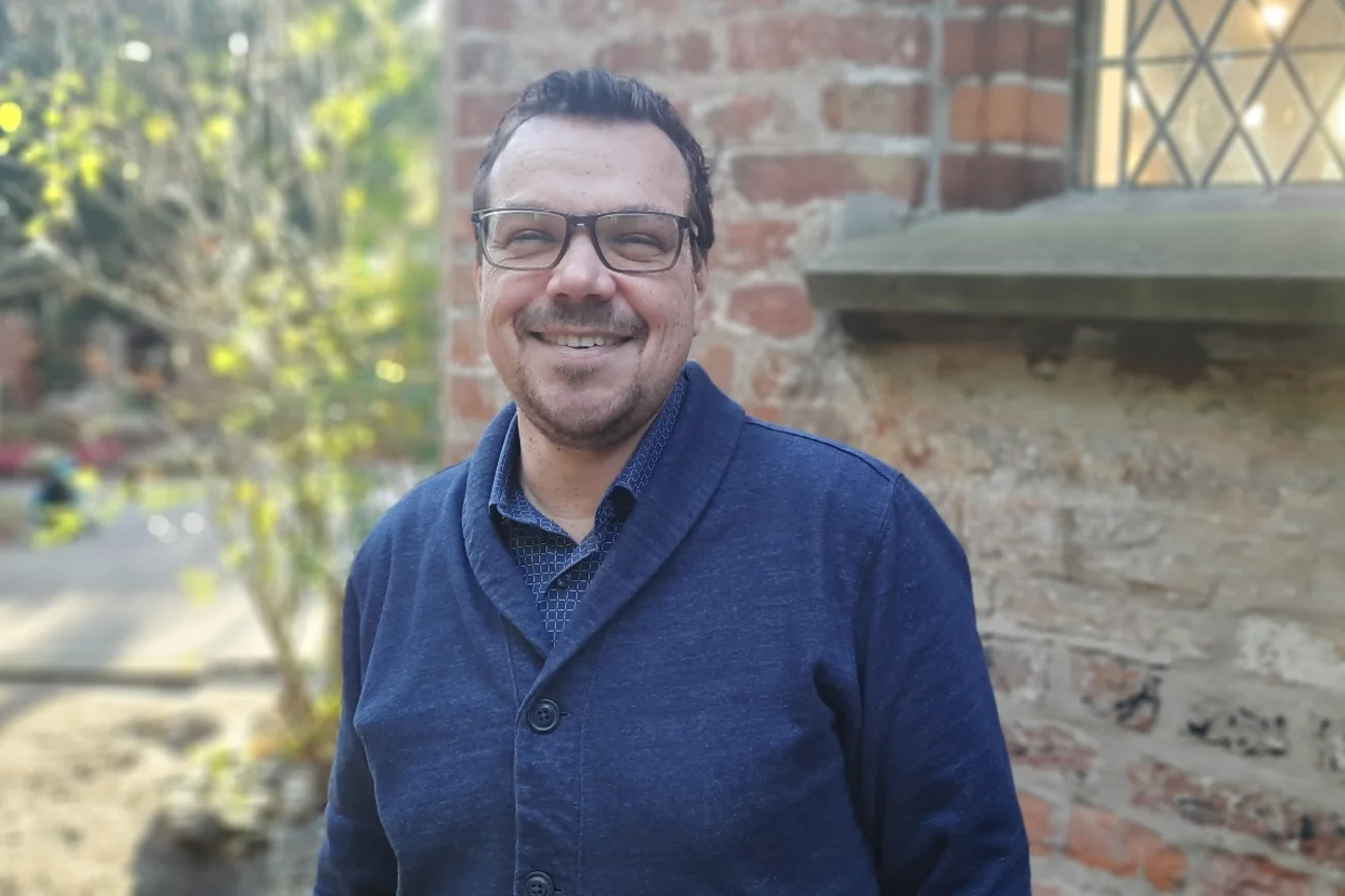 Ein Mann mit Brille & blauem Pullover schaut freundlich in die Kamera. Im Hintergrund das Mauerwerk der St. Jürgen-Kapelle.