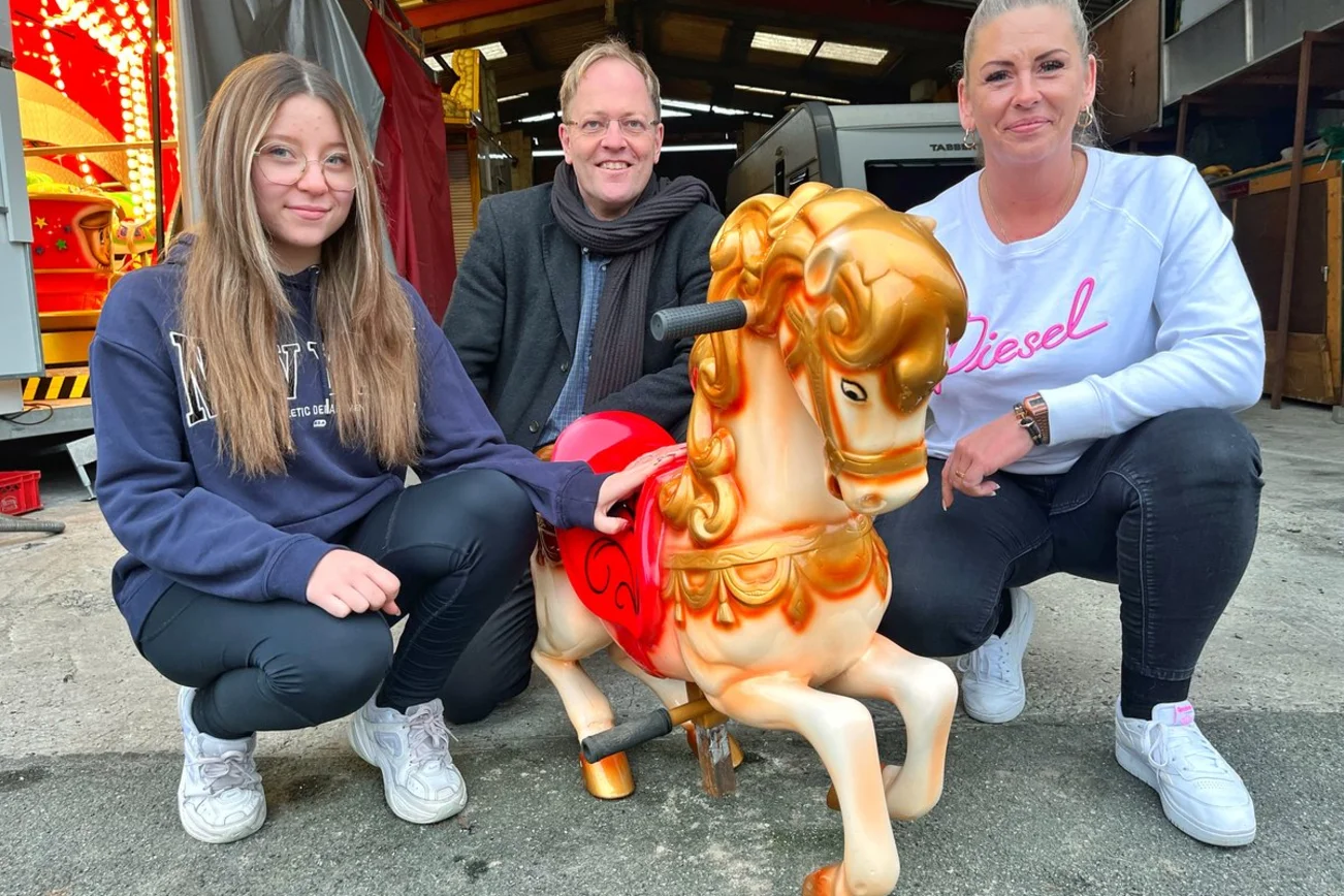 Drei Menschen stehen vor einer Lagerhalle mit einem Schaukelpferd. 
