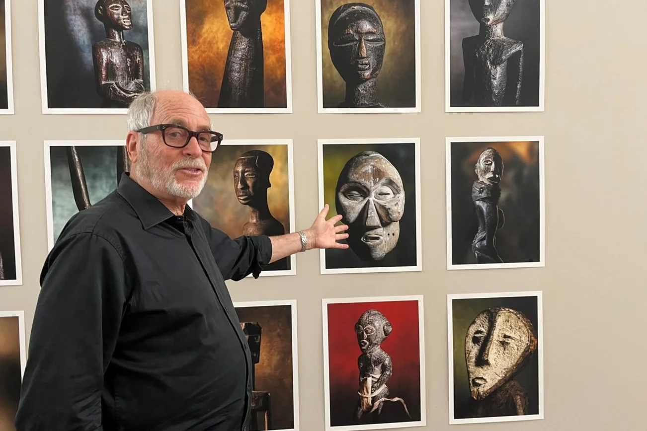Ein älterer Mann mit schwarzer Brille steht vor einer Wand mit Fotografien