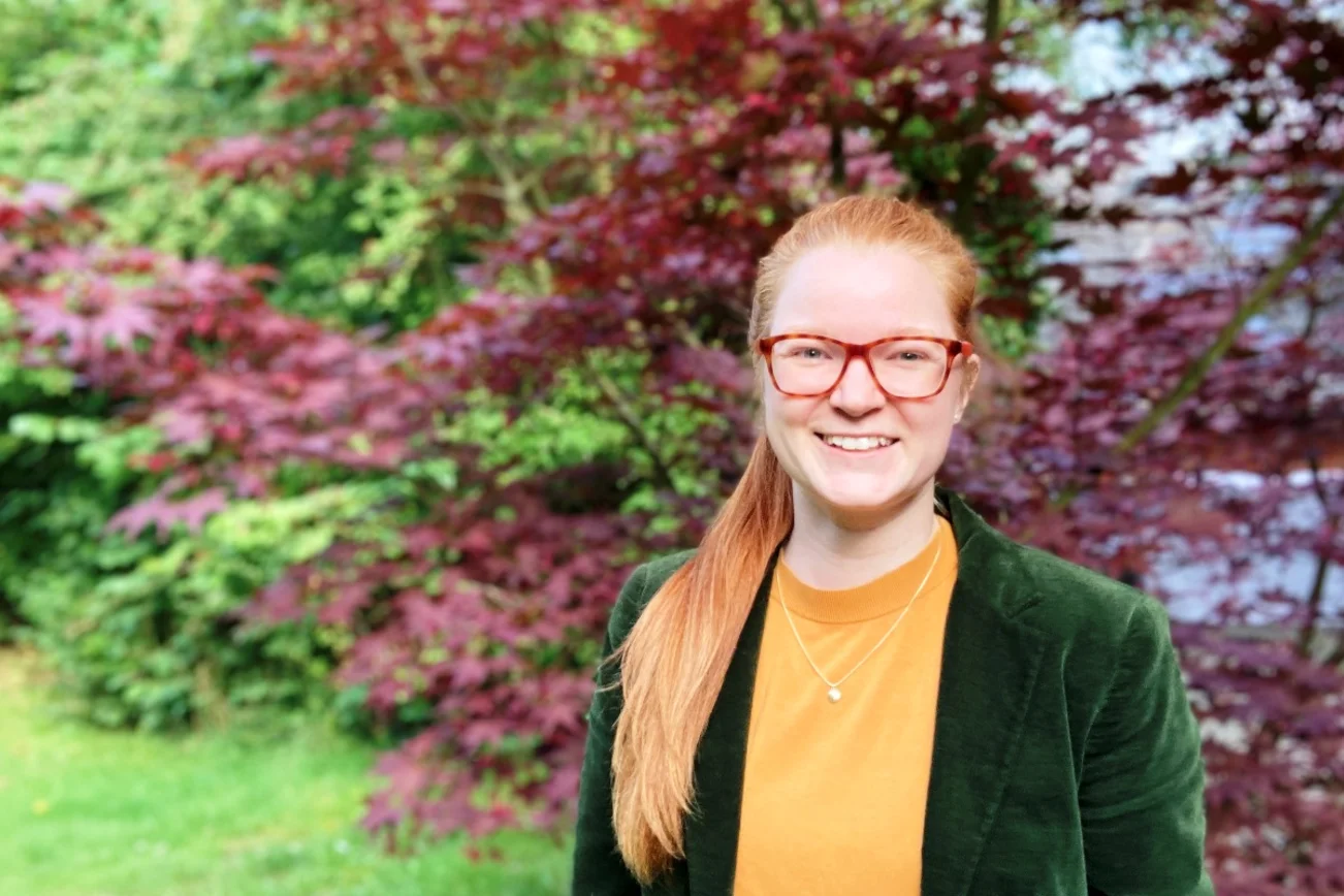 Eine Frau mit langen Haaren, Brille, grüner Jaclke und orangenem Oberteil schaut in die Kamera. Im Hintergrund eine Hecke mit grünen und roten Blättern
