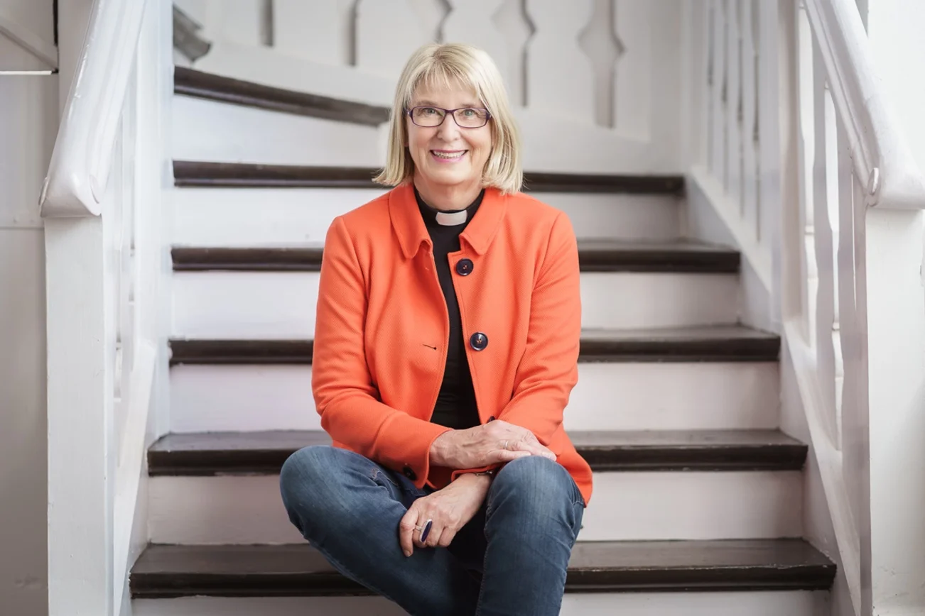 Eine Frau in Jeans, orangem Blazer und Collarhemd sitzt auf den Stufen einer weißen Holztreppe. Sie blickt freundlich in die Kamera.