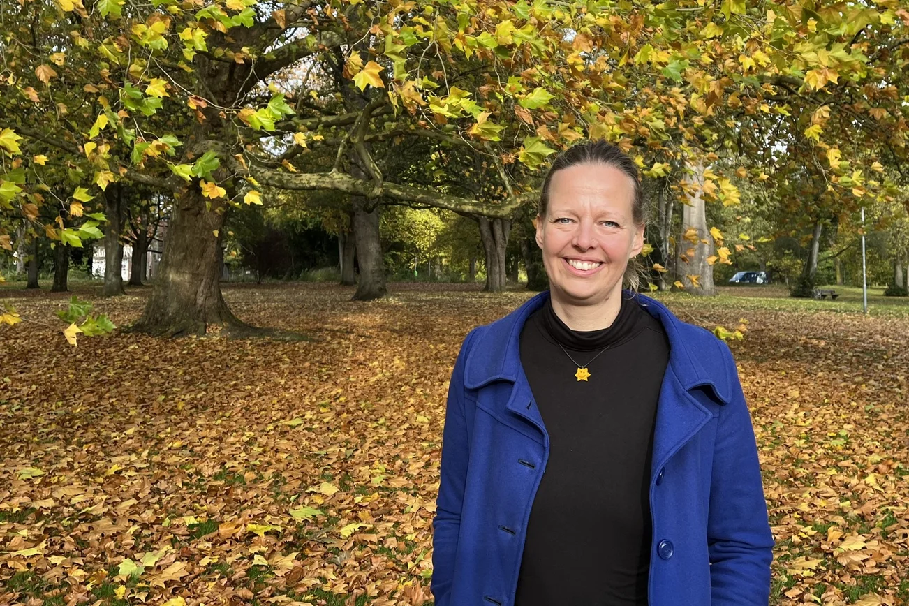 Junge Frau steht vor einem Baum und schaut lächelnd in die Kamera, viel Herbstlaub liegt um sie herum.