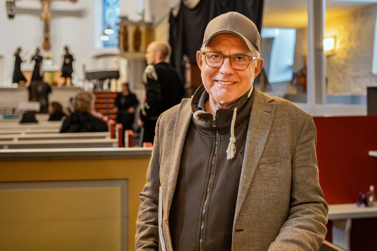 Ein Mann mit Brille, Sakko und Basecap steht in einer Kirche und lächelt in die Kamera.
