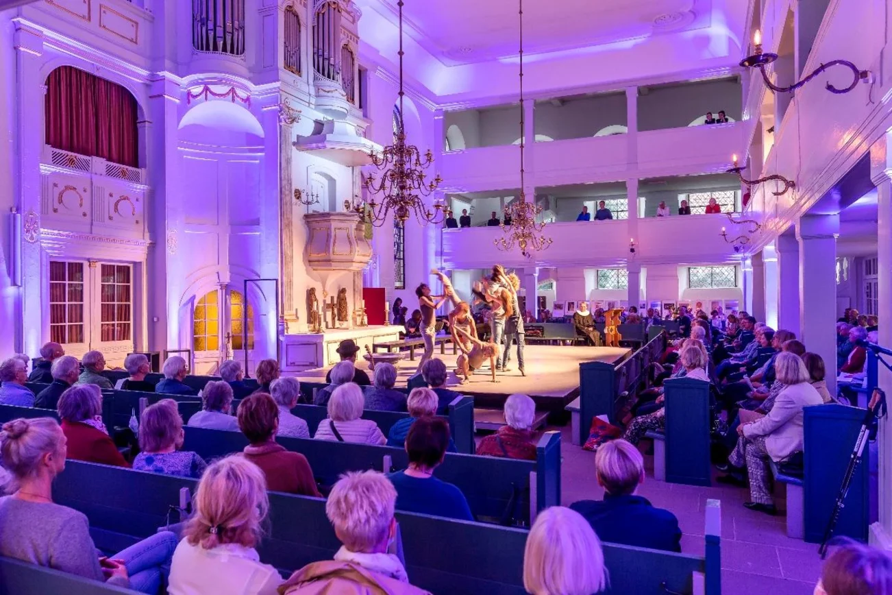 Ein Kirchraum. Vor dem Altar eine Bühne. Menschen sitzen in Reihen rund um die Bühne. Auf der Bühne Ballett.
