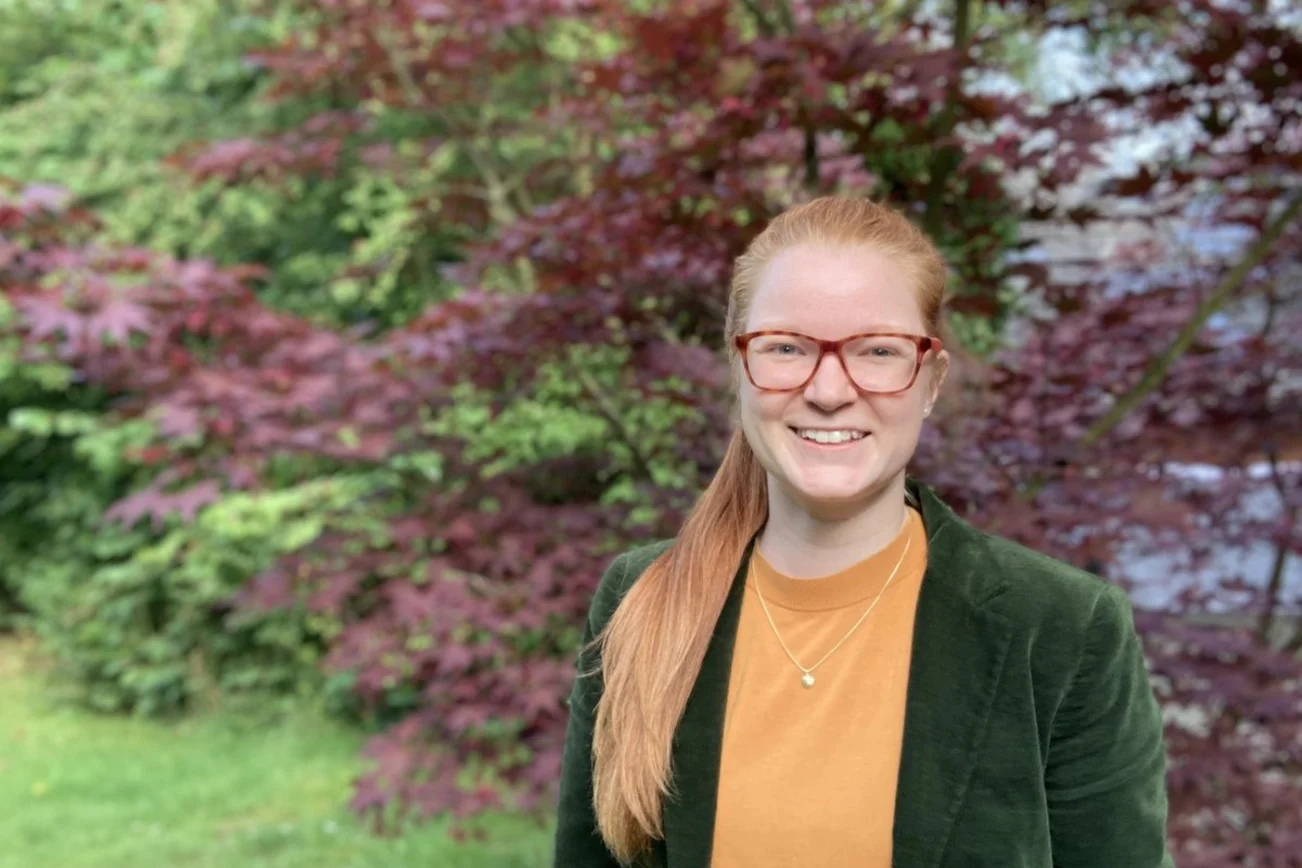 Eine Frau mit grünem Blazer, langen Haaren und Brille steht vor einem rötlichen Busch.