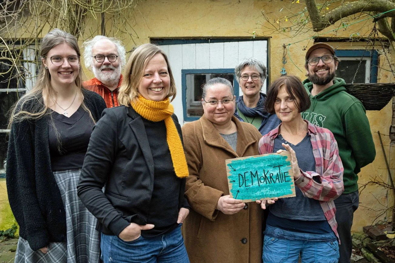 Sieben Frauen und Männer stehen als Gruppe zusammen und zeigen ein Schild mit der Aufschrift Demokratie.