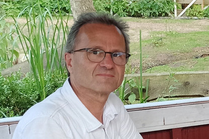 Pastor Christian Asmussen lächelt in die Kamera und trägt eine Brille. Er trägt ein weißes Hemd und im Hintergrund sieht man einen Garten mit wenig grünem Bewuchs. - Copyright: Christian Asmussen 