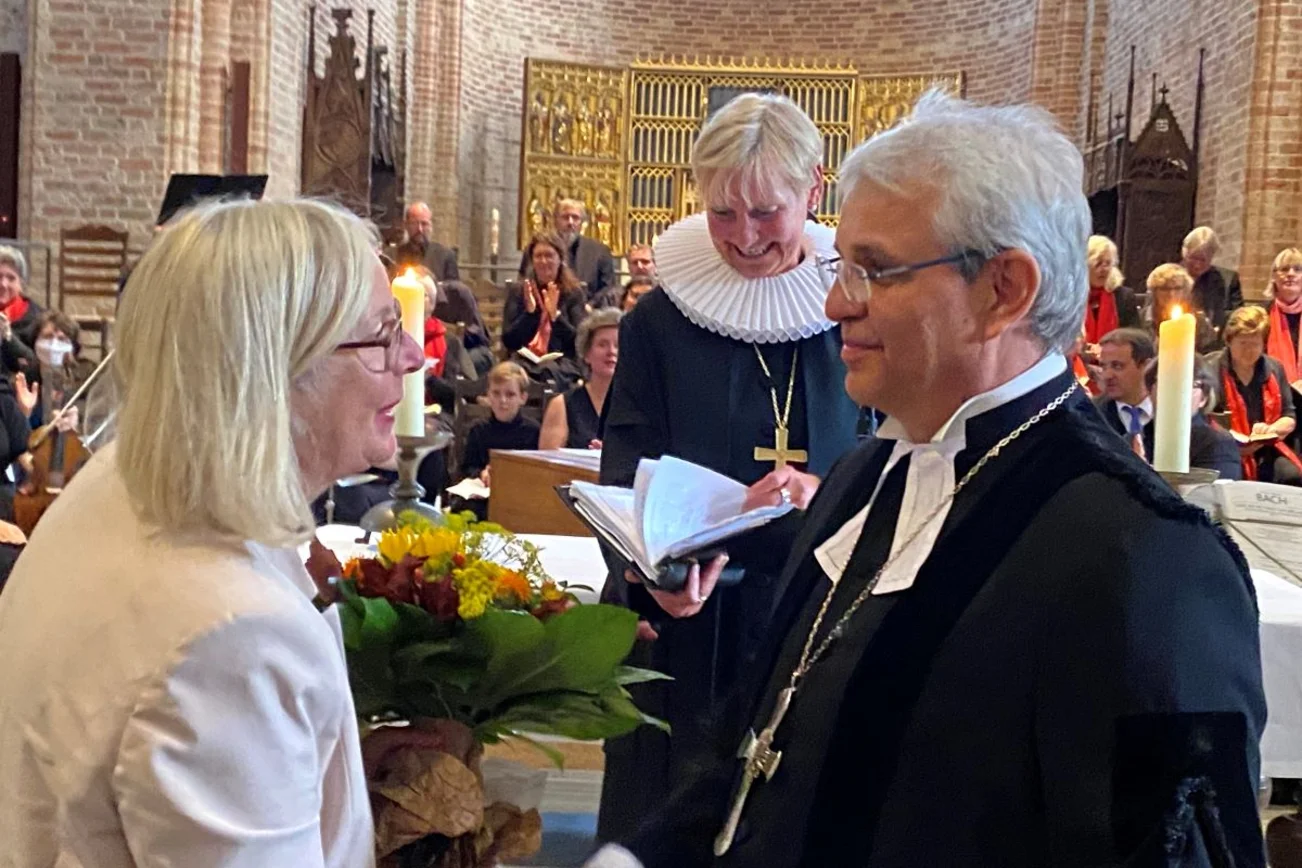 Ein Kircheraum, im Vordergrund zwei Menschen, die sich anschauen. Im Hintergrund Bischöfin, Altar mit Kerzen und Chor.