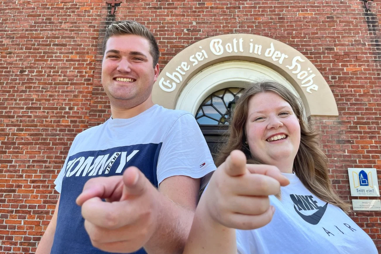 Zwei fröhliche Menschen stehen vor einer Kirche und zeigen in die Kamera