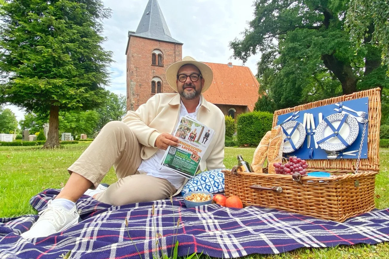 Ein mann sitzt auf einer Picknickdecke vor einer Kirche und schaut in die Kamera. 
