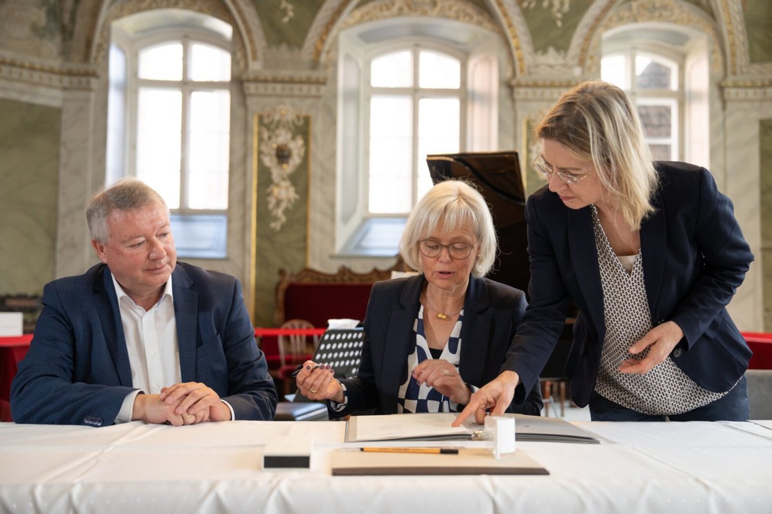 Eine Frau sitzt hinter einem Tisch, eine stehende Frau neben ihr, zeigt ihr etwas. En Mann schaut links daneben sitzend zu.