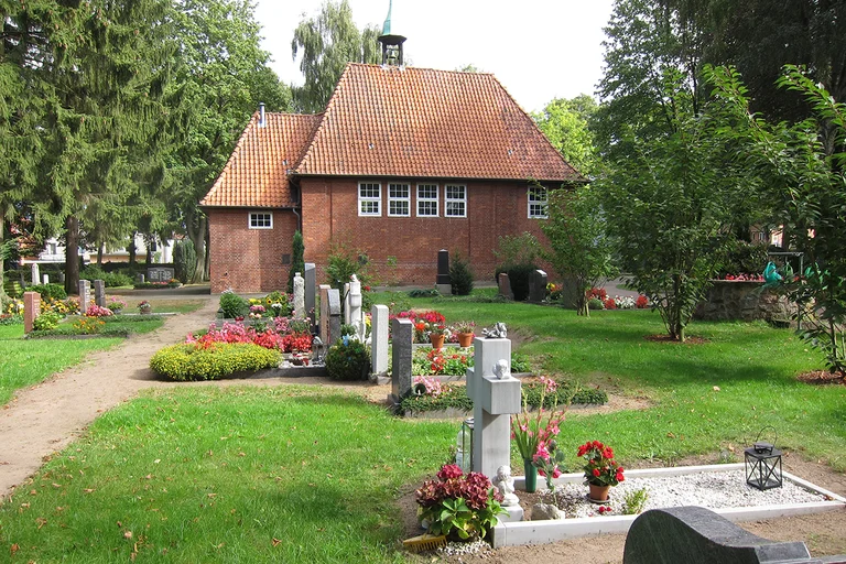 Vor der Friedhofskapelle sind mehrere Gräber, mit blühenden Pflanzen und ein Weg zu sehen