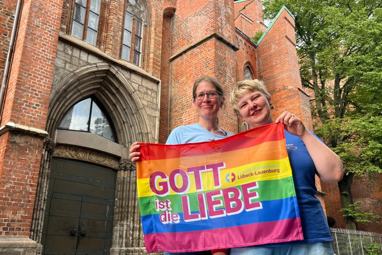 Zwei Frauen stehen vor einer Kirche und halten eine Regenbogen-Fahne mit der Aufschrift "Gott ist die Liebe"