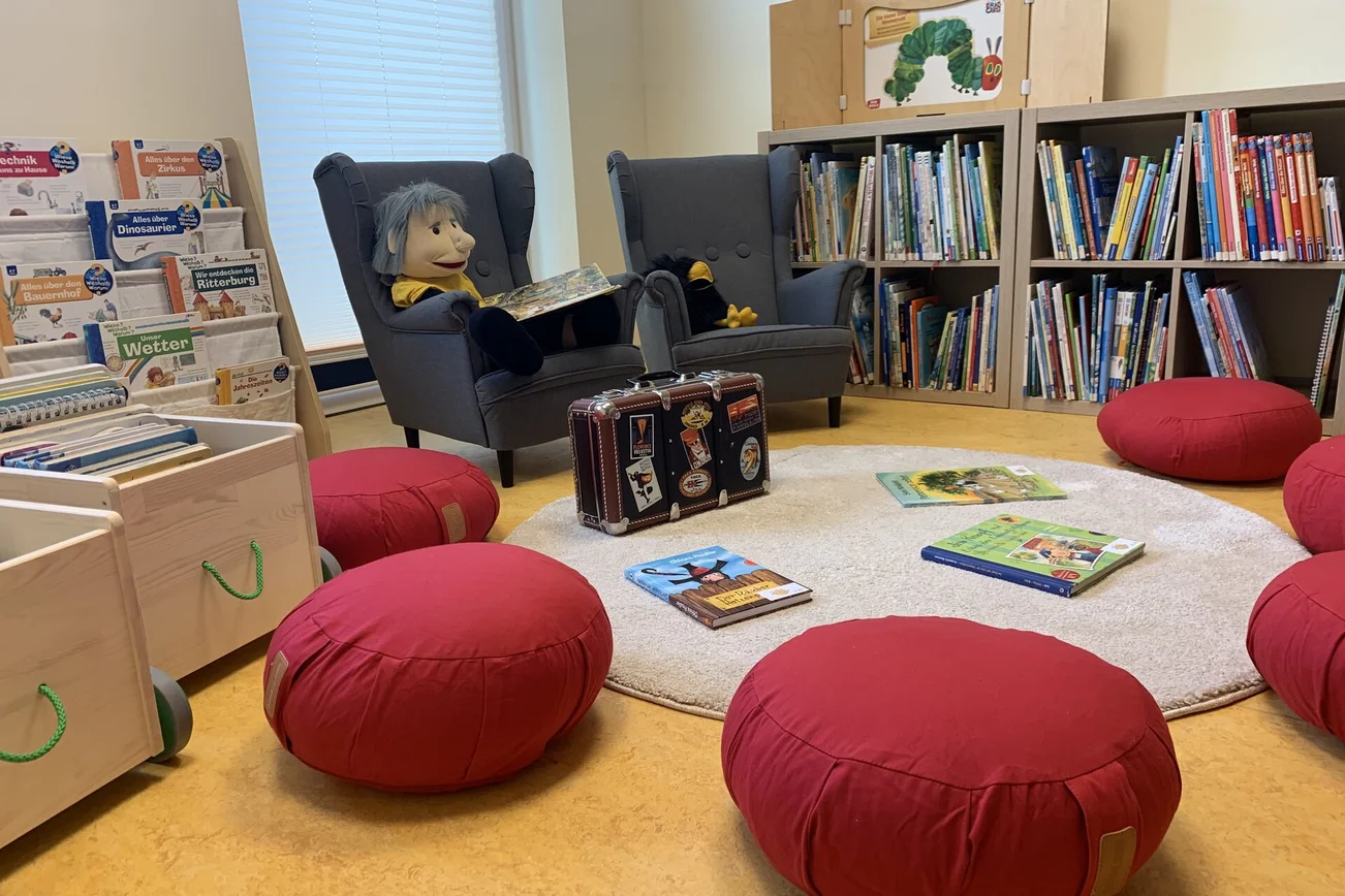Blick in eine gemütliche Kinderbücherei mit roten Sitzhockern und zwei großen Sesseln.