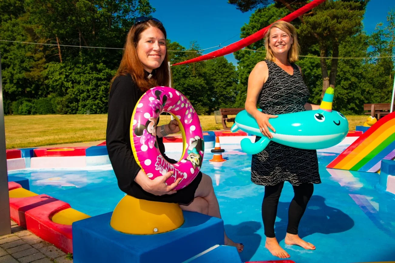 Zwei Frauen stehen an einem Schwimmbecken mit Badetieren und lächeln in die Kamera. 