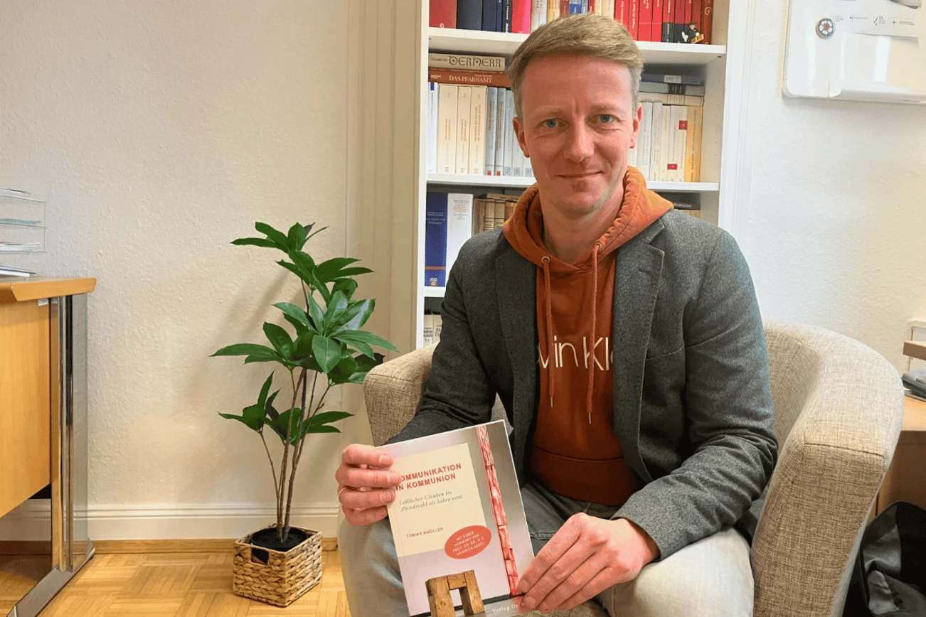 Pastor Knöller sitzt auf einem Sofa mit einem Buch in der Hand