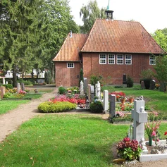 Vor der Friedhofskapelle sind mehrere Gräber, mit blühenden Pflanzen und ein Weg zu sehen