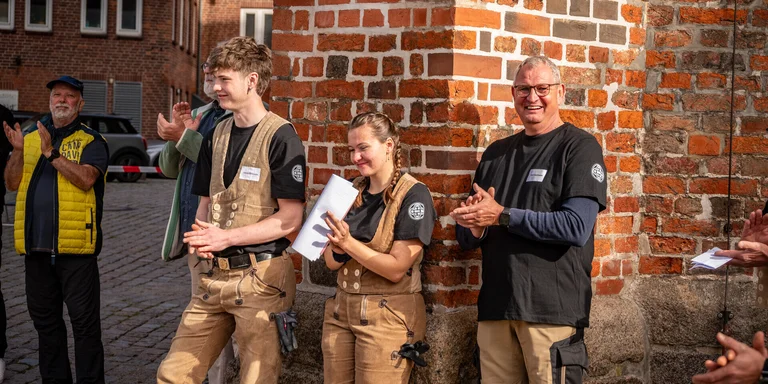 Drei Personen in Handwerkerkluft lehnen an einer Backsteinwand.