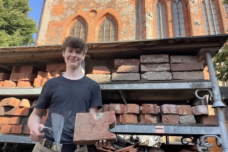 Ein junger Mann steht mit einer Maurerkelle und einem Ziegelstein vor einem Regal mit Ziegeln.