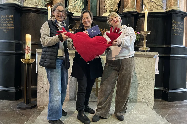 Drei Frauen stehen vor einem Altar und halten ein rotes Kuschelherz.