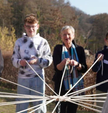 Scharf im Vordergrund ein Holzstück mit Seilen, die im Hintergrund von Personen gehalten werden.