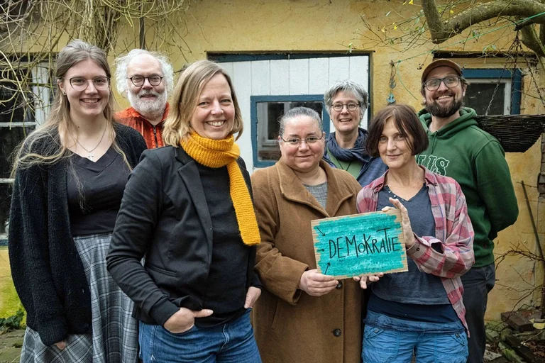 Sieben Frauen und Männer stehen als Gruppe zusammen und zeigen ein Schild mit der Aufschrift Demokratie.
