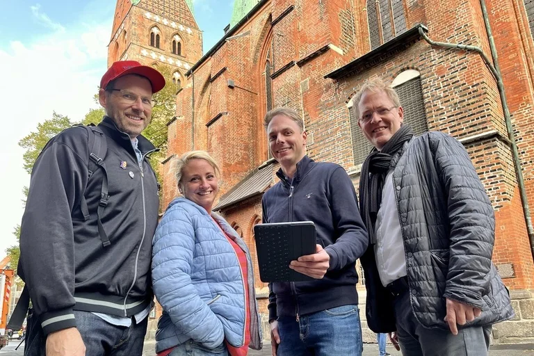 Drei Männer und eine Frau stehen vor einer Kirche und lächeln in die Kamera.
